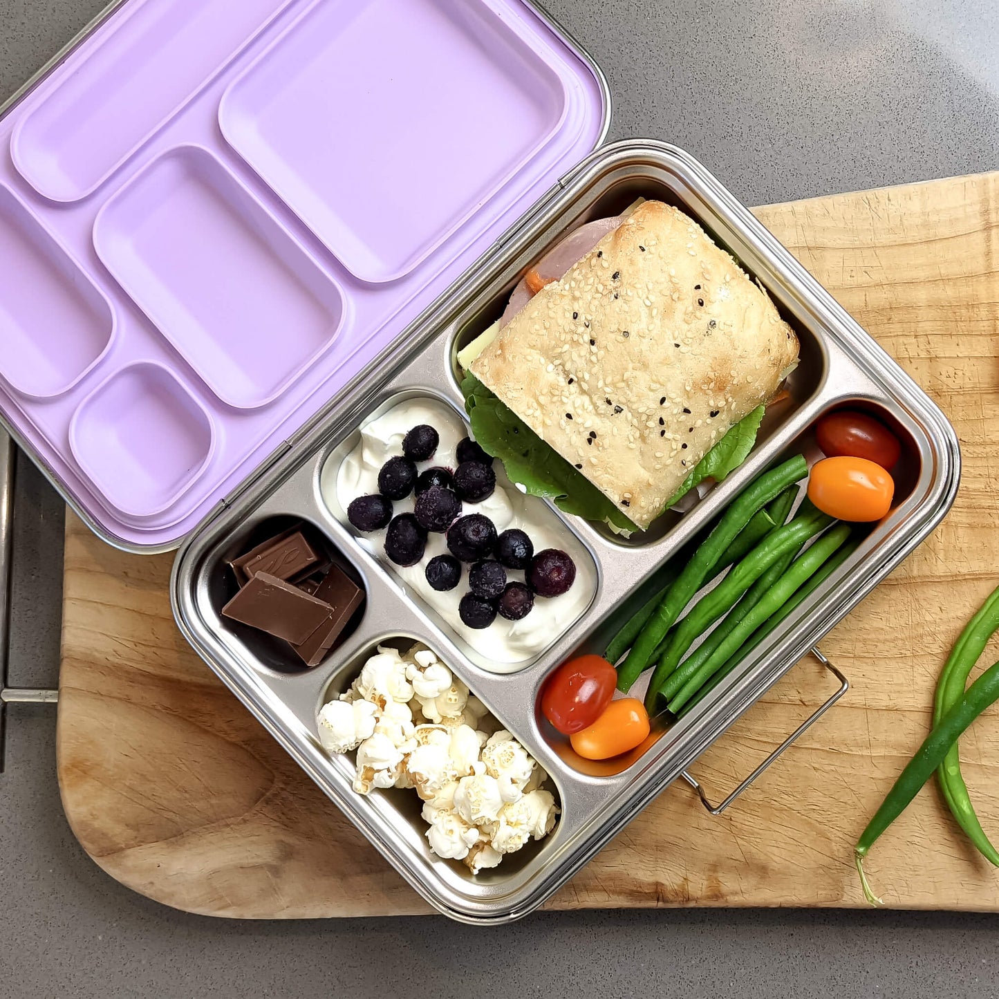 Metal lunch box with compartments containing a sandwich, fruits, vegetables, and popcorn on a wooden board.